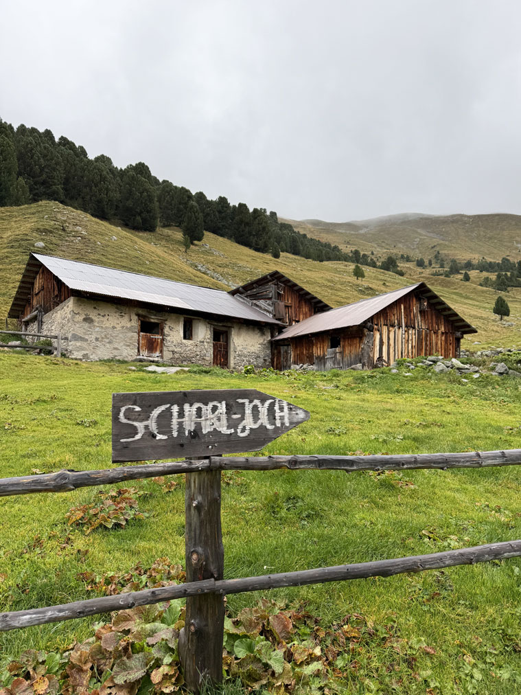 Ein hölzerner Wegweiser an einem Zaun, dahinter drei kleine Berghütten